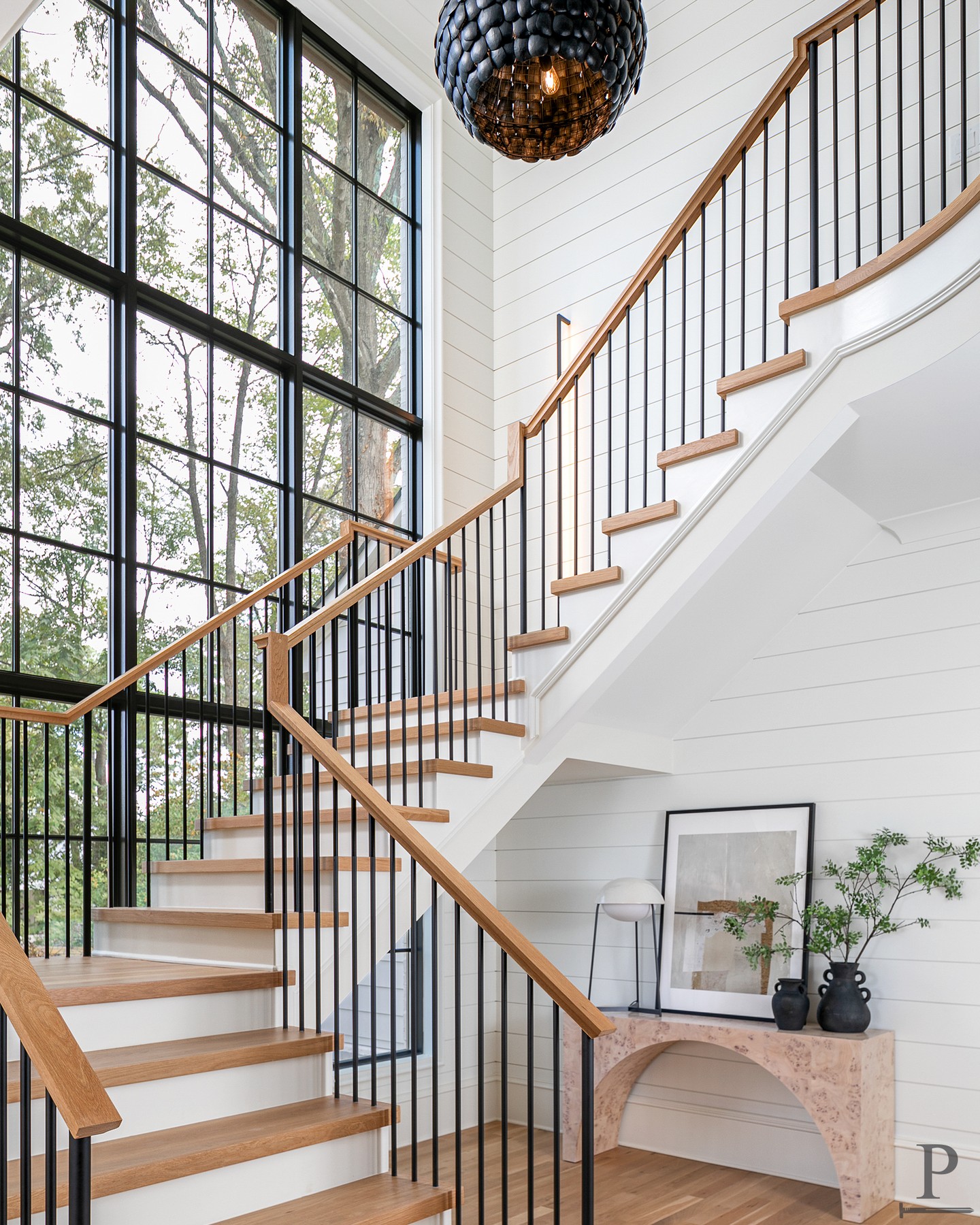The staircase is always the moment. Oak treads, black iron balusters, and a two-story steel window wall that fills every inch of the climb with light. This is what we build for.